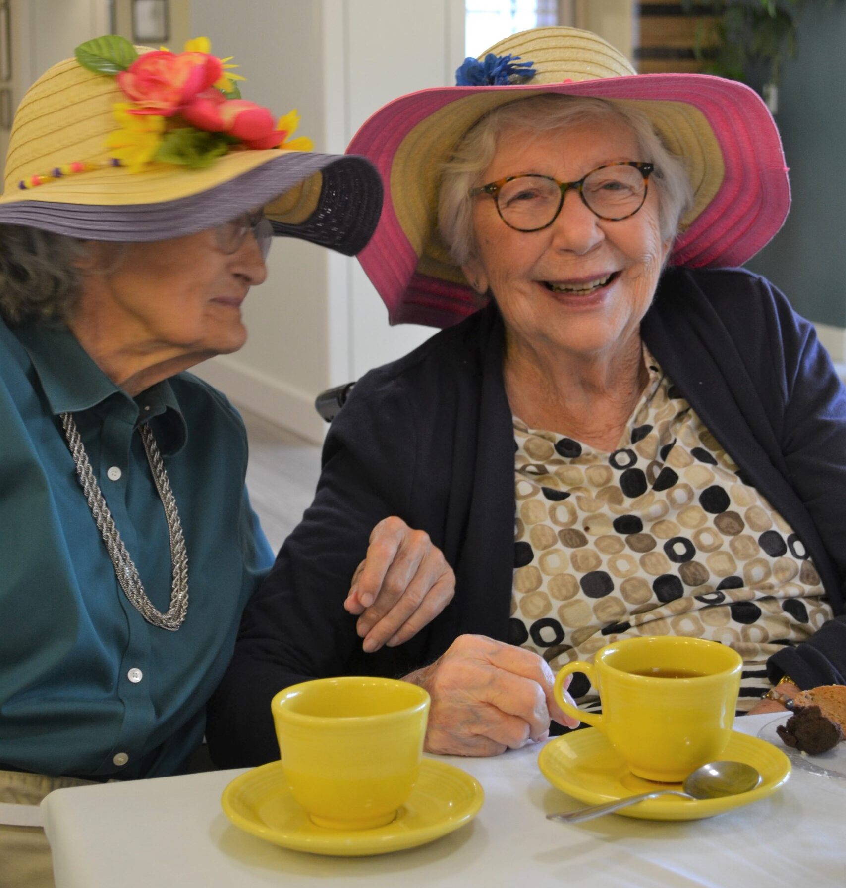 Two Memory care residents in floppy hats enjoying tea and cookies in the dining room at Abe's Garden Community